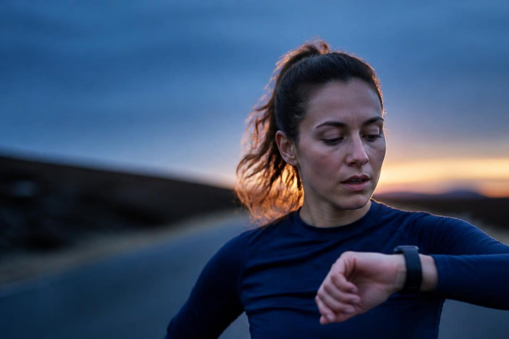 A runner at dusk, glancing down at her sports watch mid-run.